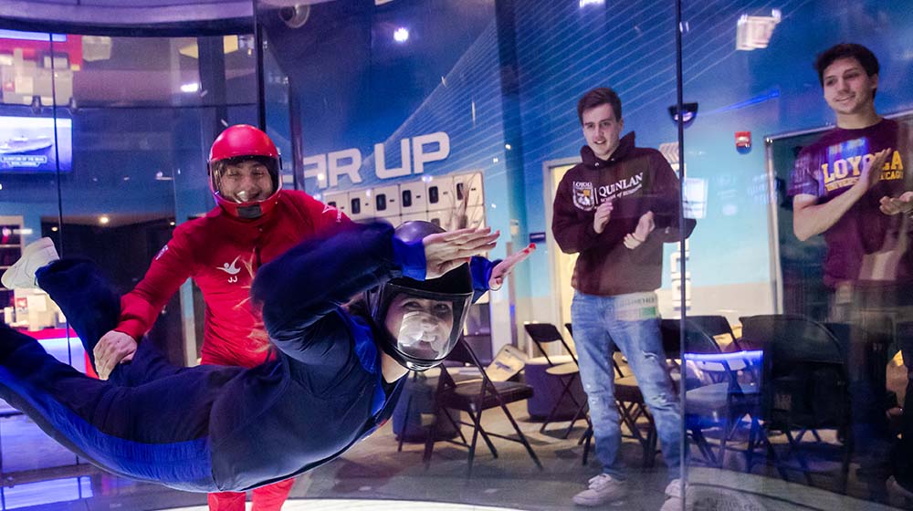A Quinlan student, smiling, indoor skydiving with an instructor while other students cheer her on
